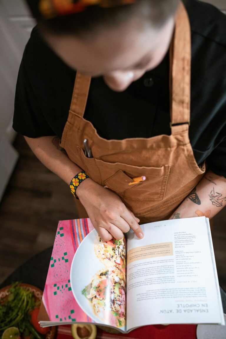 A chef in a brown apron reading a cookbook for recipe guidance while cooking indoors.